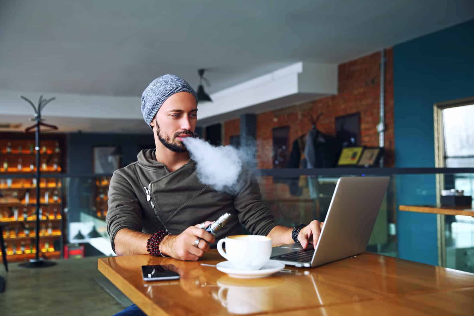 hipster man with beard sitting in cafe with a cup of coffee, vaping and releases a cloud of vapor, how to enhance your vaping experience