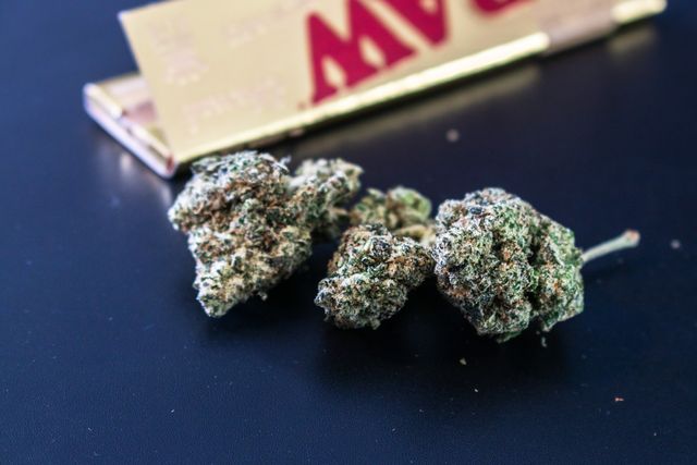 Three cannabis buds next to a RAW rolling papers booklet on a dark surface